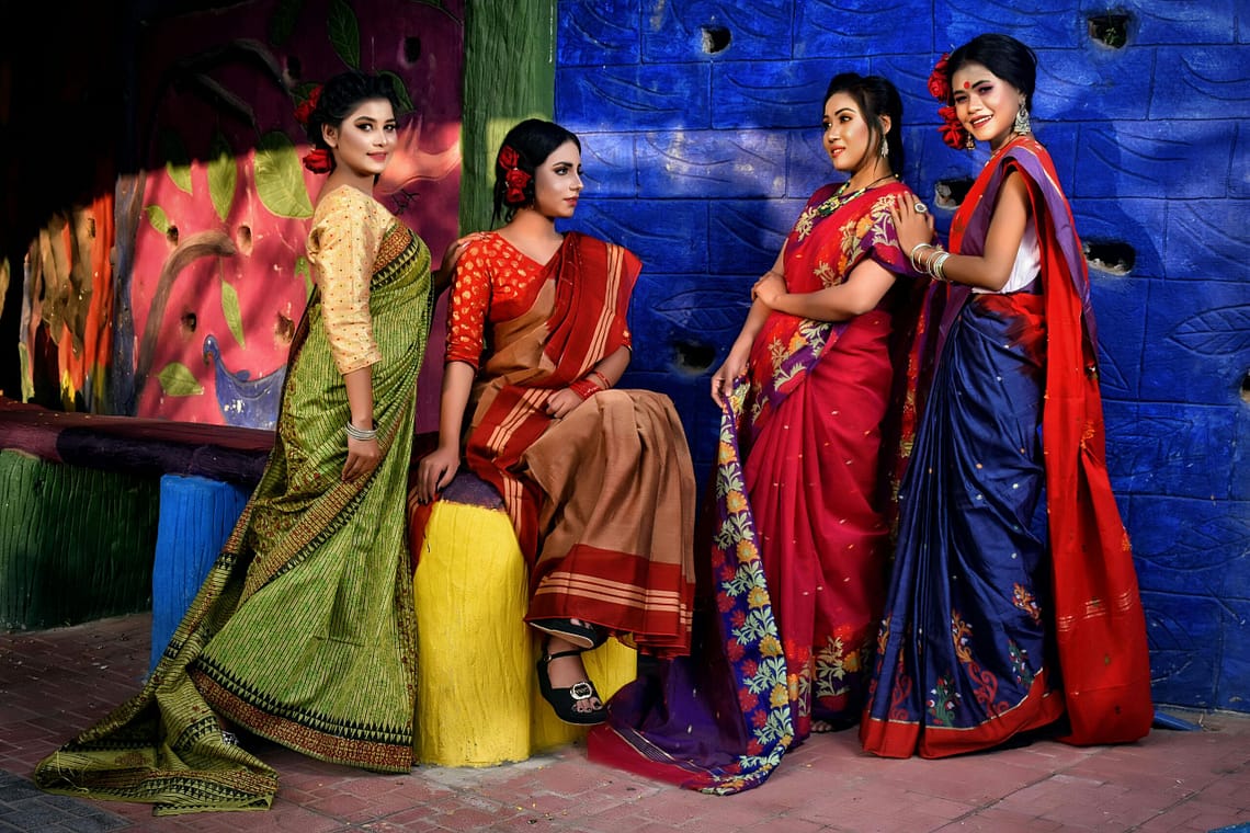 Group of women in vibrant Indian sarees posing against a colorful backdrop.