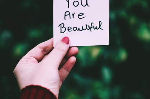 A close-up of a hand holding a 'You Are Beautiful' note against a blurred green background.