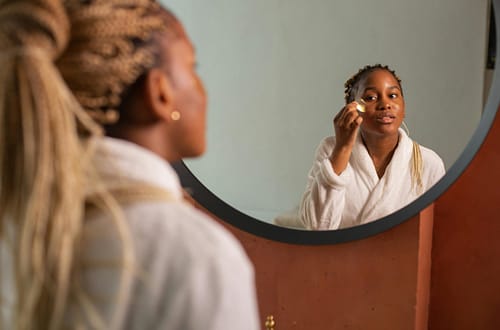 Young woman in robe applying skincare with mirror reflection, highlighting self-care and beauty routine.