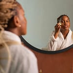 Young woman in robe applying skincare with mirror reflection, highlighting self-care and beauty routine.